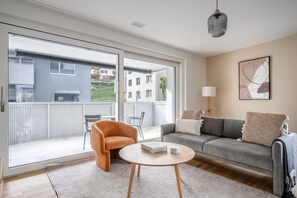 A modern living room with a grey sofa, an orange armchair, and a round coffee table. There is a sliding glass door leading to an outdoor balcony. - Hip Altstetten 1BR w/ Storage Room & W/D, nr Shops, by Blueground (Zurich)