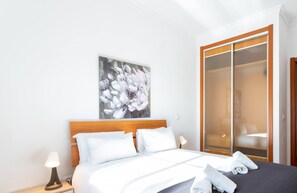 Une chambre à coucher avec un lit, une table de chevet, un tableau, une armoire et une salle de bain visible à travers une porte ouverte.