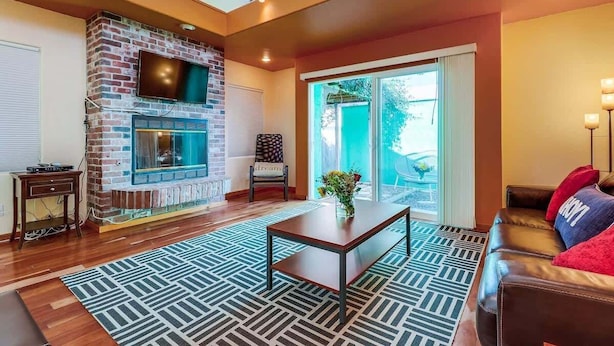 A living room with a brick fireplace, a flat-screen TV mounted above it, a sofa, a coffee table, and a patterned rug. - Discounted Rates - Ocean Beach Holiday (San Diego)