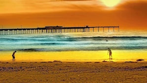 A beach with a pier extending into the water, two people walking along the shore, and the sun setting in the background. - Discounted Rates - Ocean Beach Holiday (San Diego)