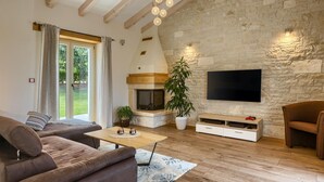 A living room with a stone fireplace, a flat-screen TV mounted on the wall, a sofa, a coffee table, and a large window with curtains. - Amazing home in Motovun (Motovun)