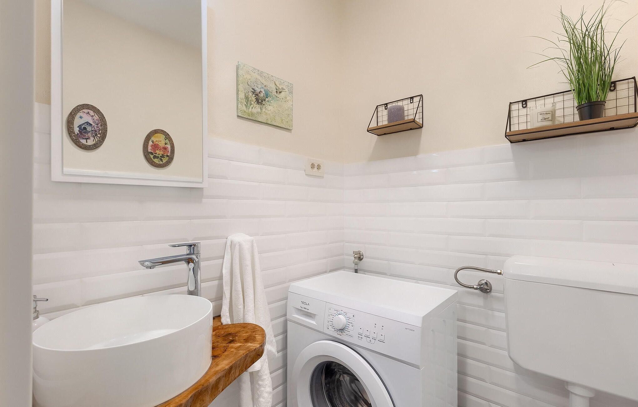 Un baño con lavamanos, lavadora e inodoro, con paredes de azulejos blancos y encimera de madera.
