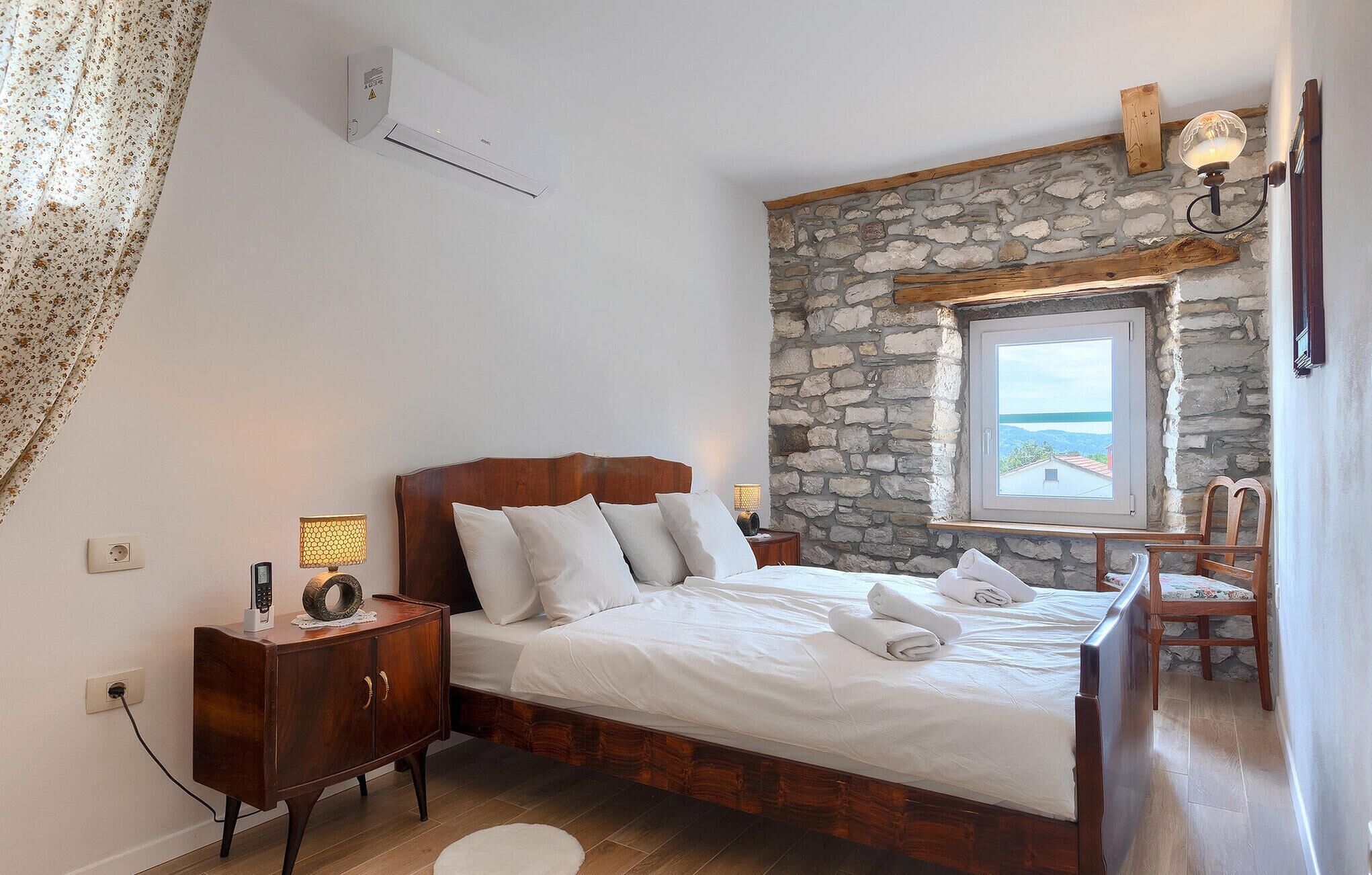Une chambre avec un mur en pierre, un lit recouvert de draps blancs, une table de chevet en bois, une lampe et une vue sur la mer par la fenêtre.