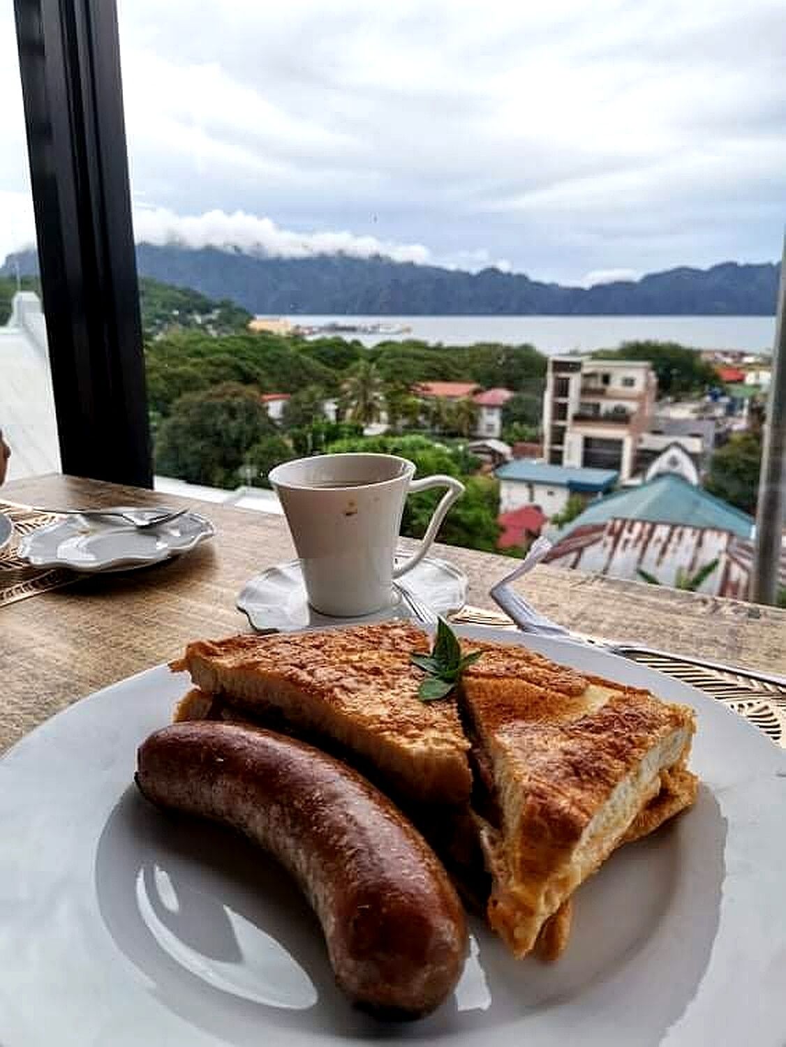A meal of grilled sandwich and sausage on a plate, with a cup of coffee and a scenic view of a town and mountains in the background.