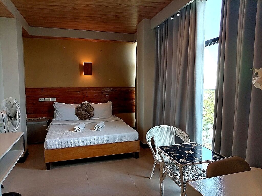 A hotel room with a bed, bedside table, chair, and a small table with a fan.