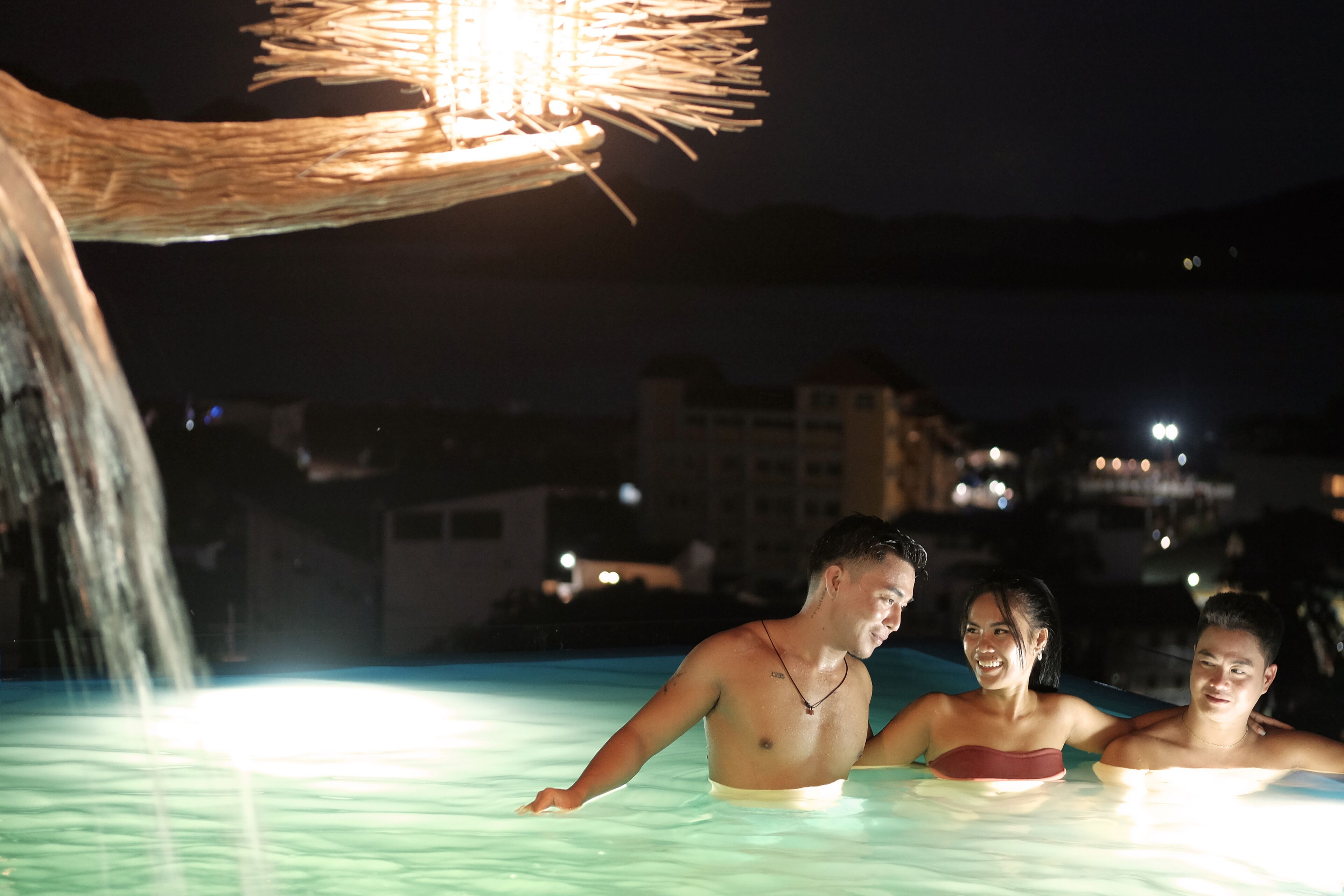 A nighttime swimming pool with a water feature and a thatched roof overhead.