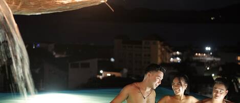 A nighttime swimming pool with a water feature and a thatched roof overhead.
