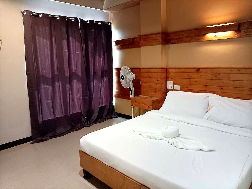 A hotel room with a wooden headboard, a bed with white linens, a fan, and a curtain.