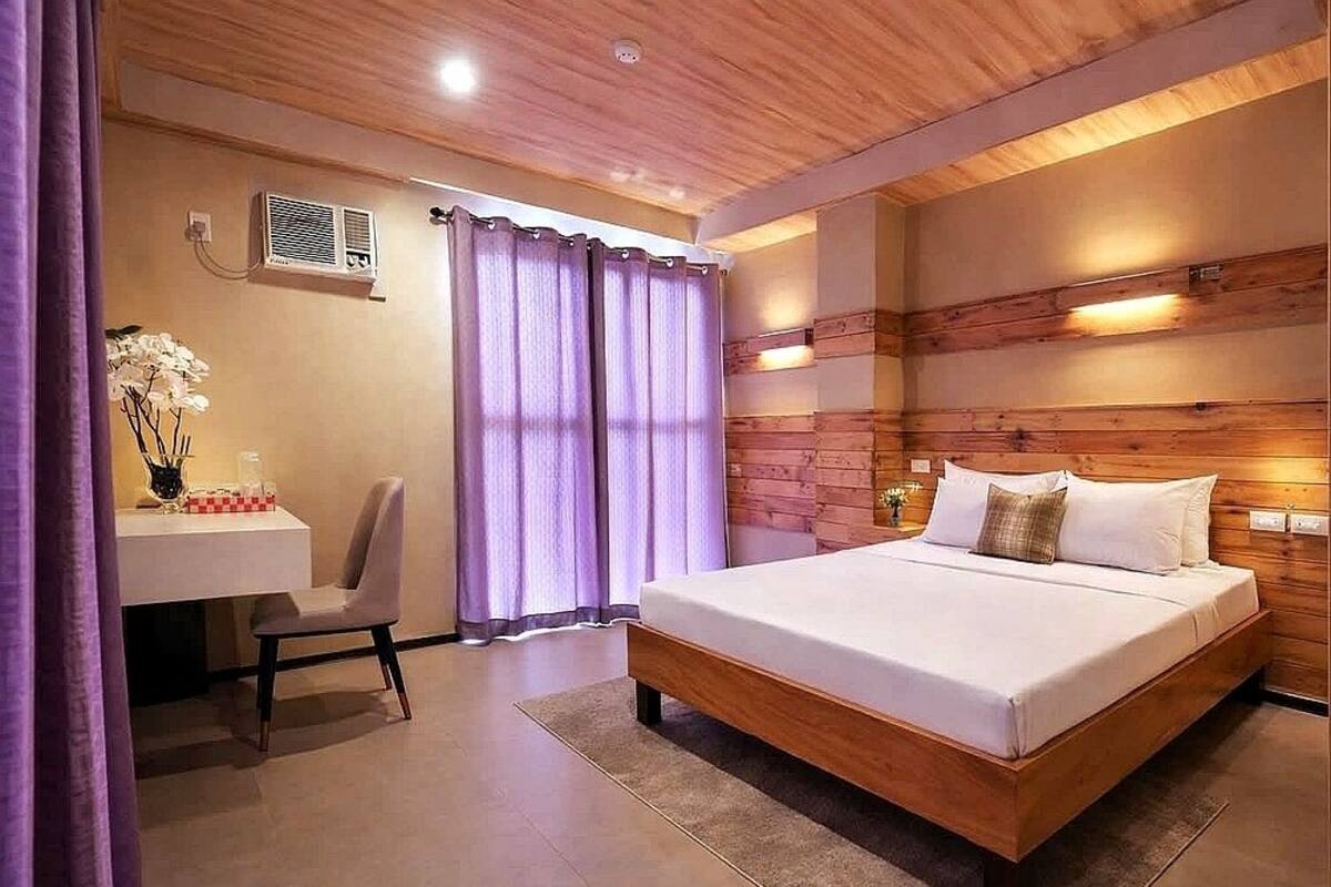 A bedroom with a wooden headboard, a bed with pillows, a desk with a chair, and a window with curtains.