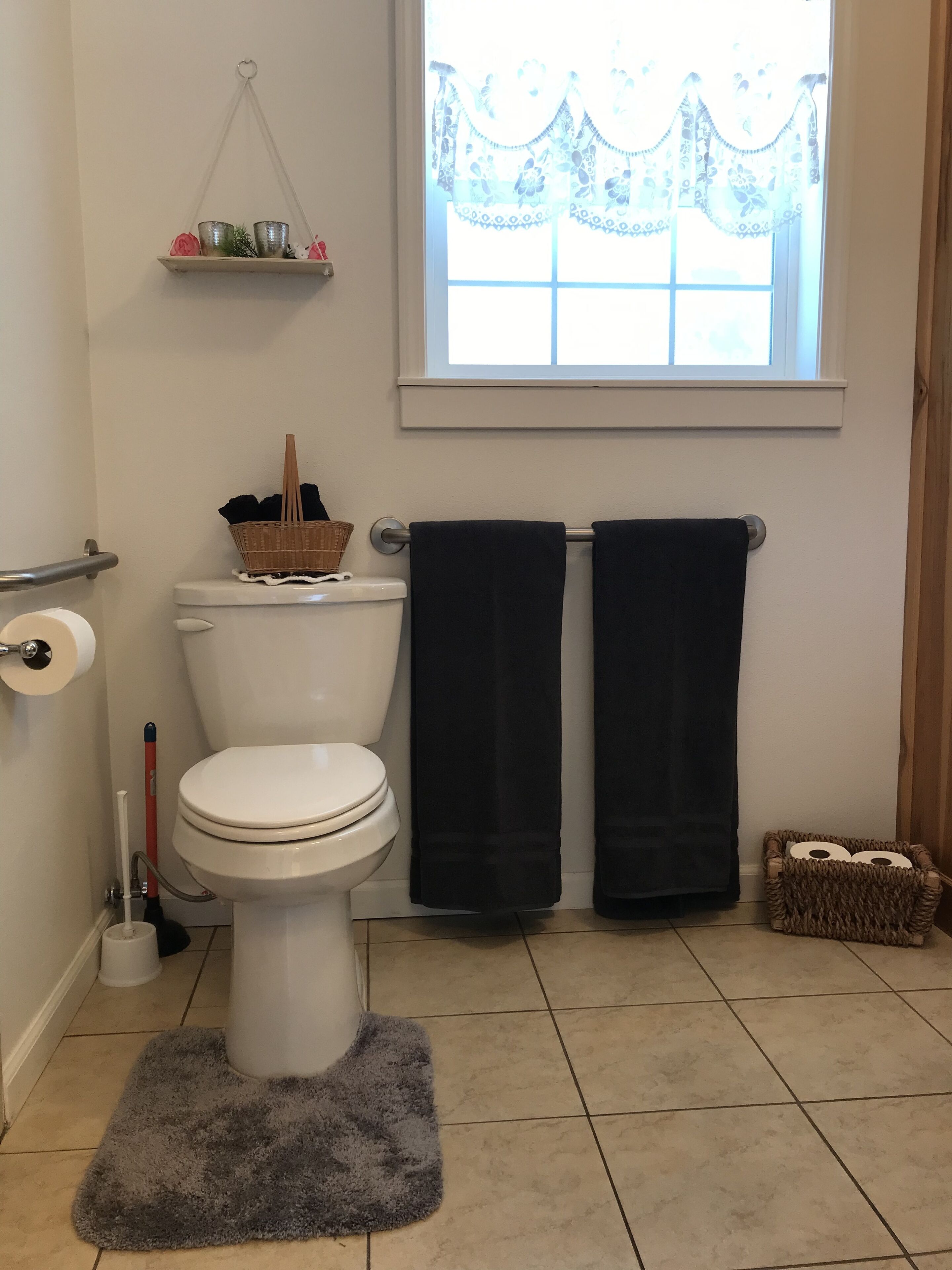 A bathroom with a toilet, a roll of toilet paper, a black towel rack, a window with a curtain, and a small shelf with decorative items.