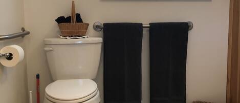 A bathroom with a toilet, a roll of toilet paper, a black towel rack, a window with a curtain, and a small shelf with decorative items.