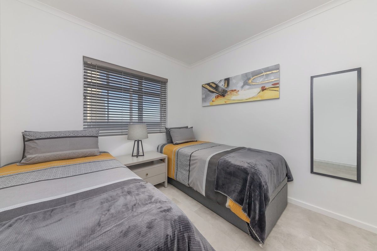 A bedroom with two beds, a window with blinds, a bedside table with a lamp, and a wall-mounted mirror.