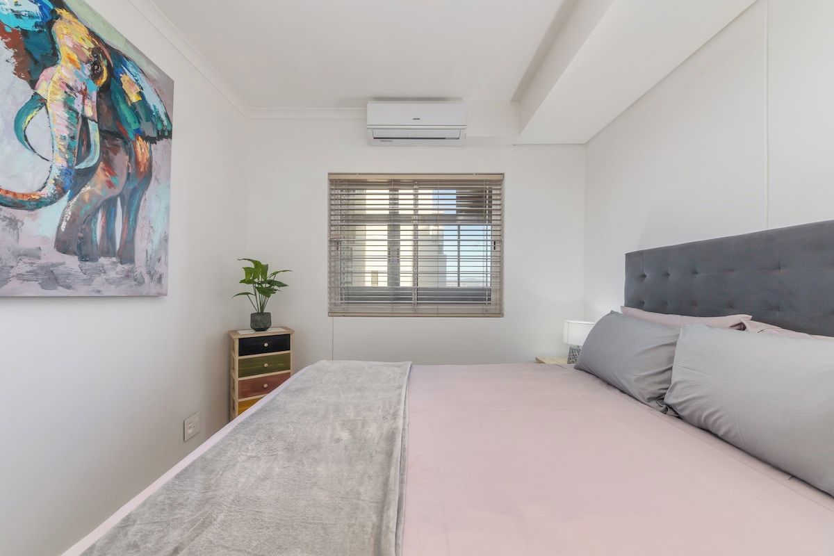Une chambre à coucher avec un lit, une fenêtre avec des stores, un climatiseur fixé au mur et un tableau coloré.
