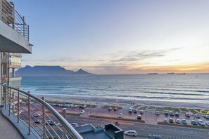 A coastal view from a balcony overlooking a parking area with cars, a beach, and ships in the distance. - 3-Bed apartment with Sunset Sea Views, Blouberg (Cape Town)