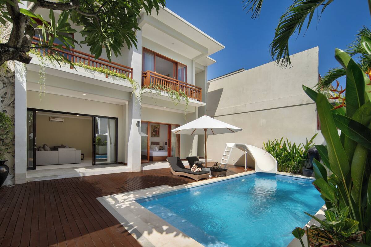 A modern hotel with a pool, a wooden deck, and a balcony with plants.