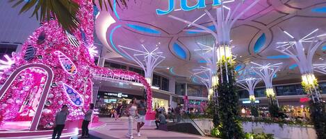A shopping mall interior with a prominent 'JUNOS' sign, decorative pink and blue lighting, and a modern ceiling design.