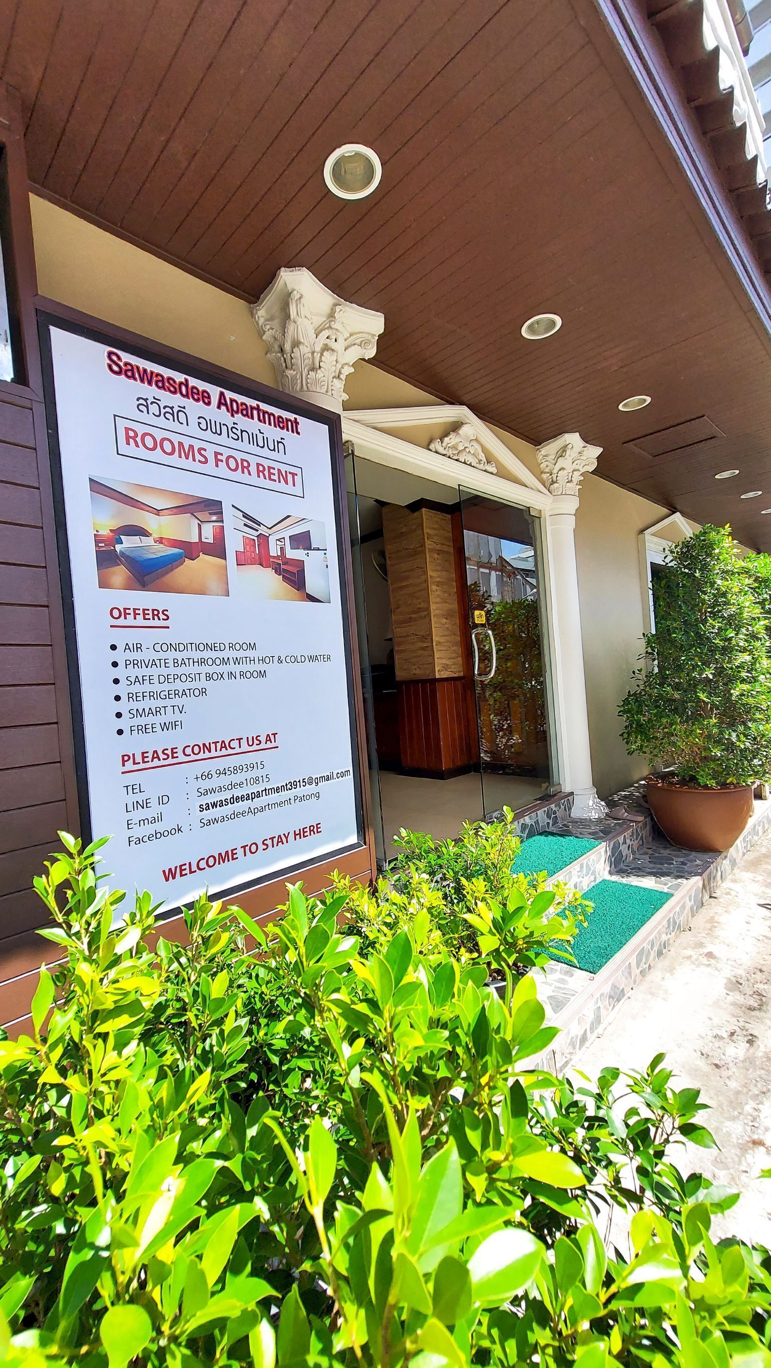 A signboard for Sawasdee Apartment detailing amenities like air-conditioned rooms, private bathroom, safe deposit box, refrigerator, smart TV, and free WiFi.