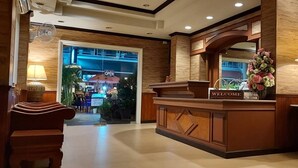 A reception area with a wooden reception desk, a 'WELCOME' sign, a large vase of flowers, and a lamp on a side table. - Sawasdee Apartment Patong (Patong)