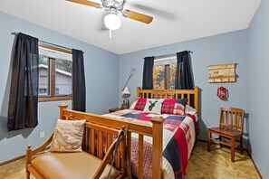 A bedroom with a wooden bed, a chair, a nightstand, a lamp, a ceiling fan, and a window with curtains.