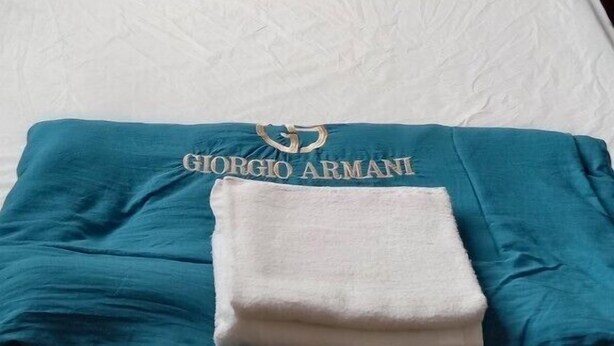 Una cama bien hecha con sábanas blancas, una manta azul con el logotipo de 'GIORGIO ARMANI' y un cabecero de madera.