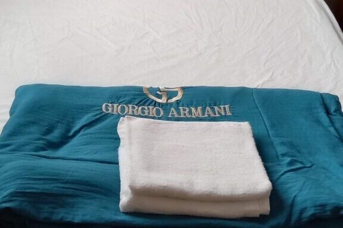 A neatly made bed with white linens, a blue blanket with "GIORGIO ARMANI" branding, and a wooden headboard.