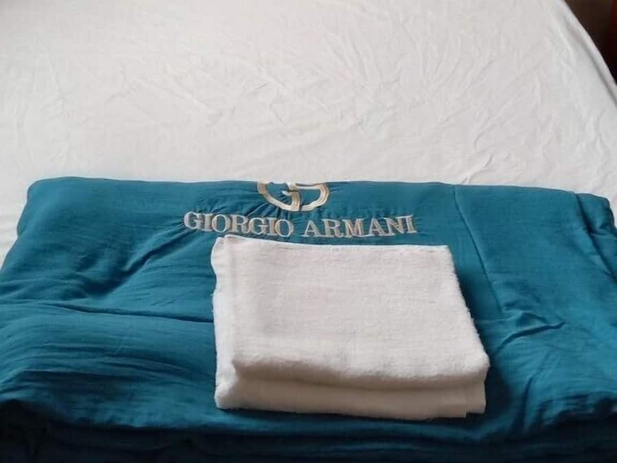 Ein ordentlich bezogenes Bett mit weißen Leinen, einer blauen Decke mit "GIORGIO ARMANI" Schriftzug und einem Holzkopfende.