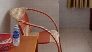 Habitación de esquina con una sola silla, una mesa con una botella de agua y una cortina.