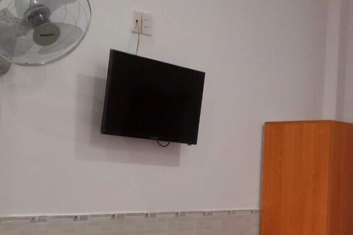 A room with a television mounted on the wall, a wooden wardrobe, a fan, and a pair of slippers on the floor.