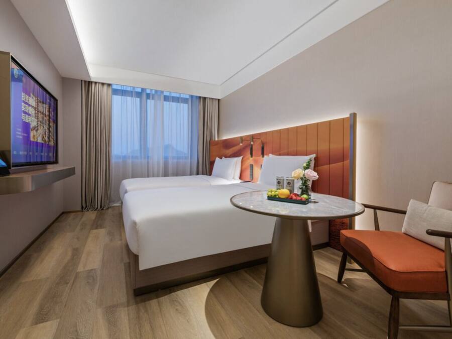 A modern hotel room with a large bed, a round table with fruits, a sofa, and a TV on the wall.