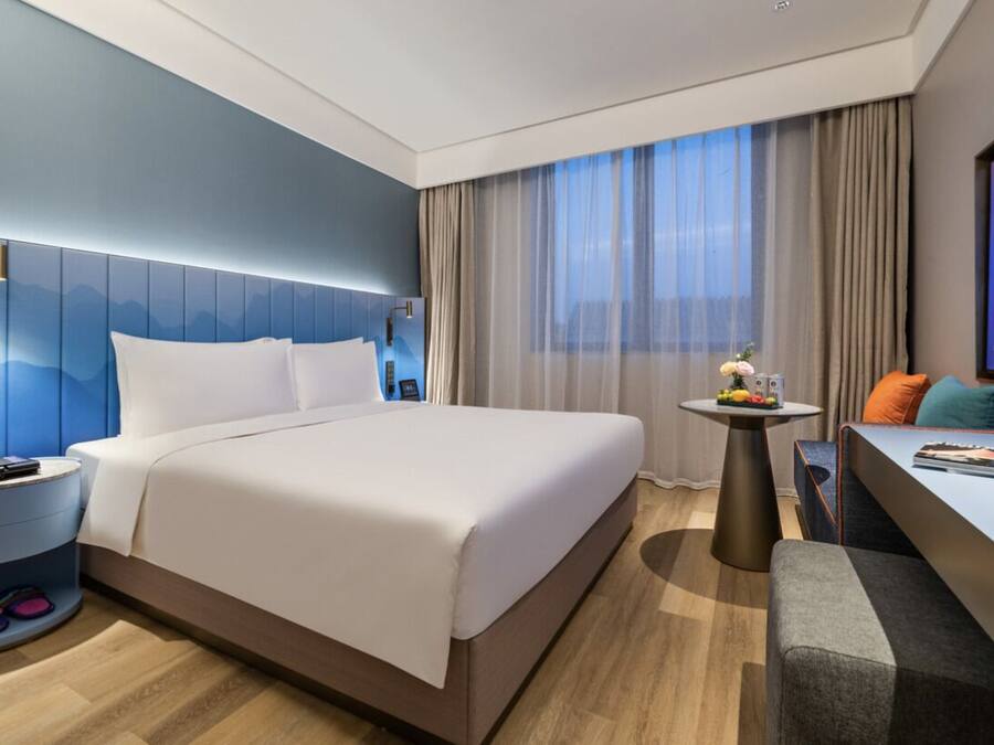 A modern hotel room with a large bed, a small side table, a seating area with a sofa, and a TV on the wall.
