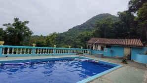A swimming pool with a blue tiled deck, a blue building with a tiled roof, and a mountainous backdrop. - Chalé Mirante do Félix Ubatuba  (Ubatuba)