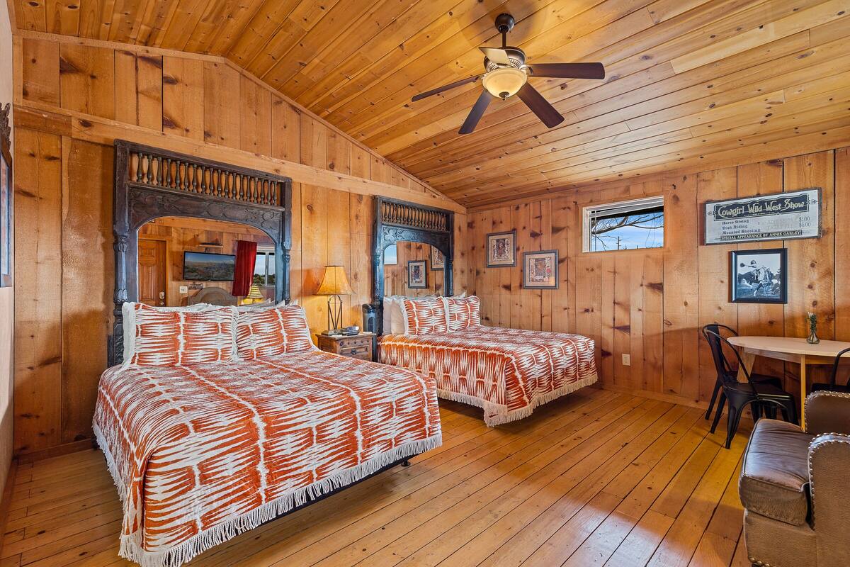 Une pièce de style cabane en bois, avec deux lits, un ventilateur de plafond, une table à manger avec des chaises et des tableaux encadrés accrochés aux murs.