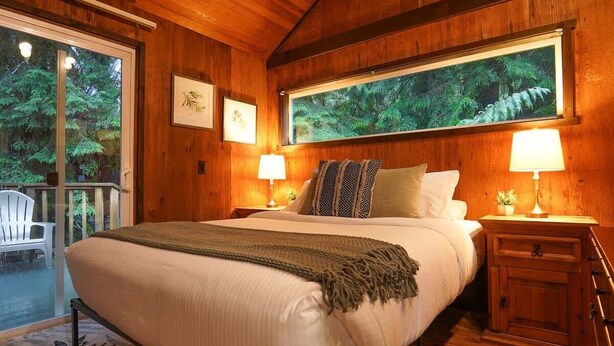 Una habitación de madera con una cama, dos mesitas de noche, un ventanal con vistas a un bosque y una silla afuera.