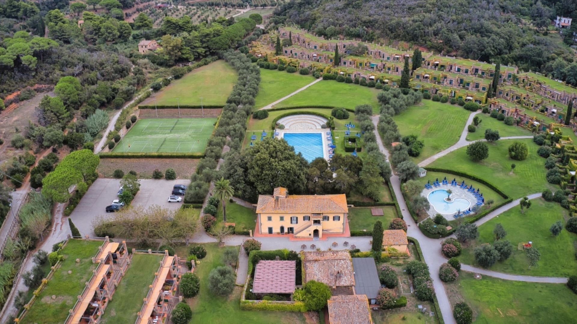 An aerial view of a large estate with a central building, tennis courts, a swimming pool, and extensive gardens.
