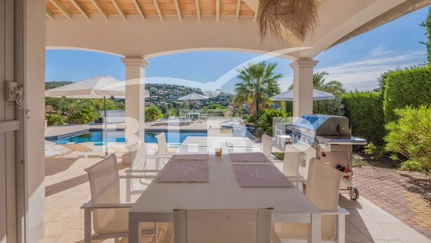 A covered outdoor dining area with a pool view, white furniture, and a grill. - Modern villa in the prestigious golf domain (STE MAXIME)