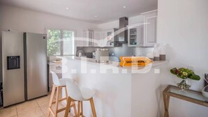 A modern kitchen with white cabinets, a stainless steel refrigerator, and a central island with bar stools. - Modern villa in the prestigious golf domain (STE MAXIME)