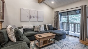 A modern living room with a sectional sofa, a wooden coffee table, and a large window with a view of trees.