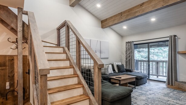 A staircase with wooden steps and a metal railing leading to a living area with a sofa, a coffee table, and a view of the outdoors through a glass door.