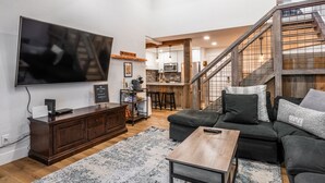 A modern living room with a large flat-screen TV, dark wooden furniture, a staircase, and a kitchen in the background.