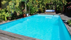 A rectangular swimming pool with a wooden deck in the foreground, surrounded by various tropical plants and a covered seating area in the background.