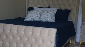 A bedroom with a tufted headboard, a bed with dark bedding, a clock on the wall, and a window with curtains. - Luisandro (Punta Cana)