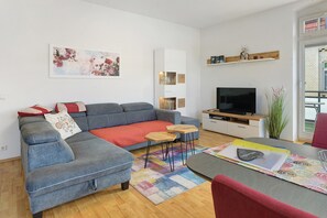 Moderni olohuone, jossa on harmaa kulmasohva, litteänäyttöinen televisio, pieni puinen pöytä ja värikäs matto.