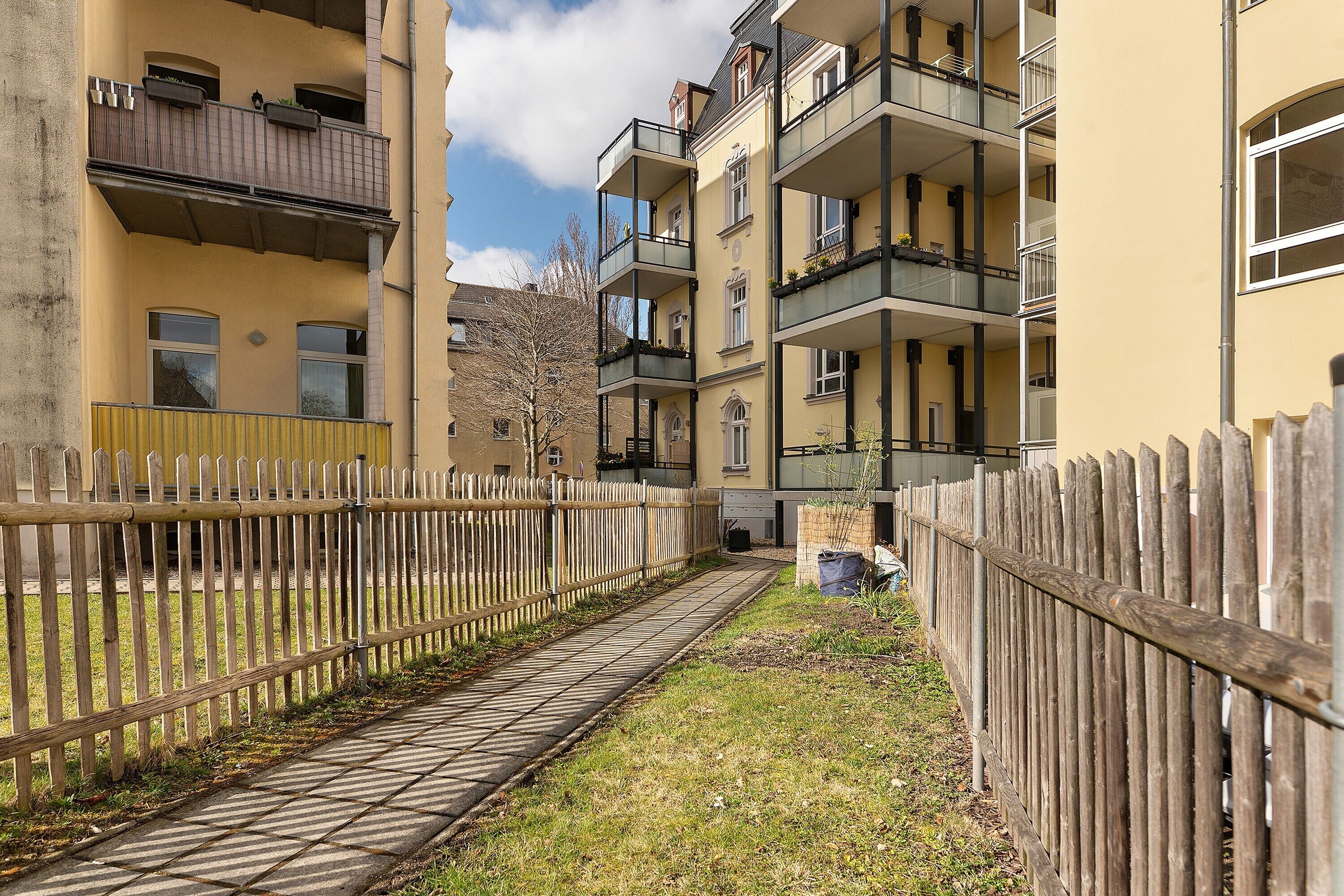 A multi-story residential building with balconies, a wooden fence, and a paved pathway.