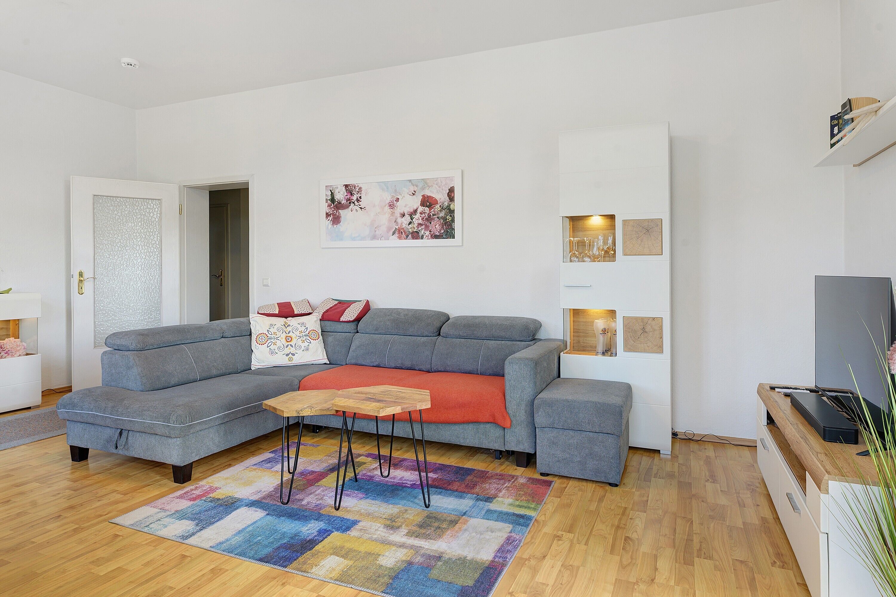 Una sala de estar con un sofá gris, una pequeña mesa de madera, una alfombra colorida y un televisor.