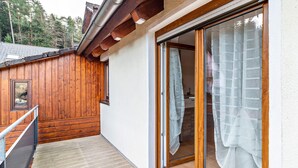 Ein Gebäude mit Holzfassade, einer Schiebeglastür, einem kleinen Balkon mit Metallgeländer und Blick auf Bäume im Hintergrund.