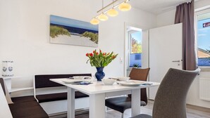 Ein Essbereich mit einem weißen Tisch, schwarz-weißen Bänken, einer Vase mit roten Blumen und einem Strandbild an der Wand.