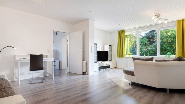 Ein modernes Wohnzimmer mit einem weißen Sofa, einem Esstisch, einem Fernseher und einem großen Fenster mit Vorhängen.