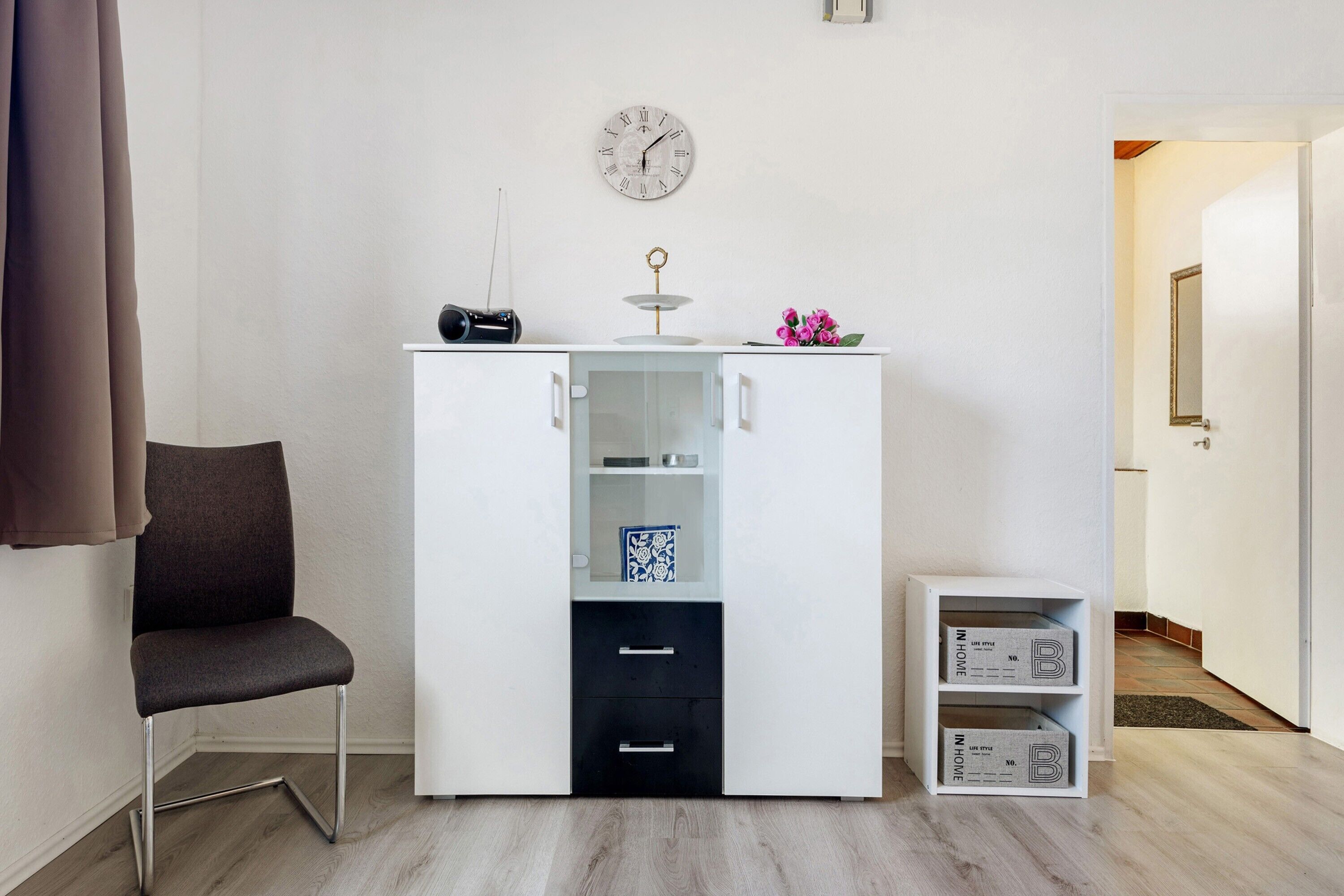 Een kamer met een witte kast, een stoel, een klok aan de muur en een deur die naar een andere kamer leidt.