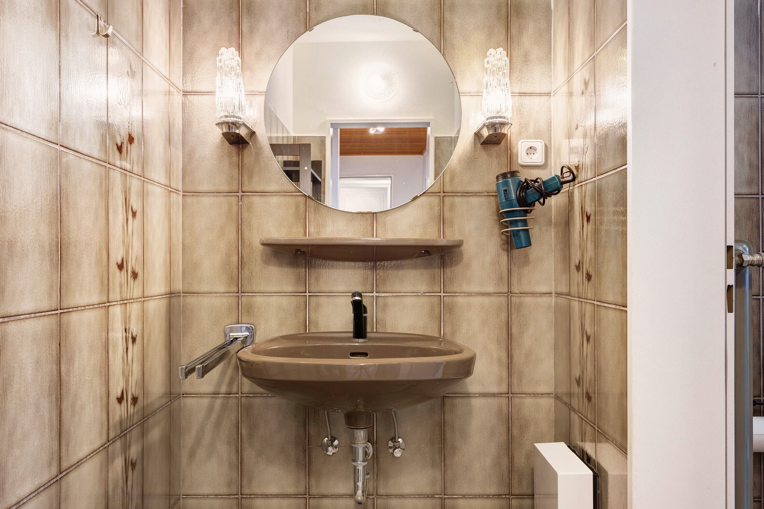 A bathroom with a wooden panel wall, a round mirror, a sink with a faucet, and two wall-mounted light fixtures.