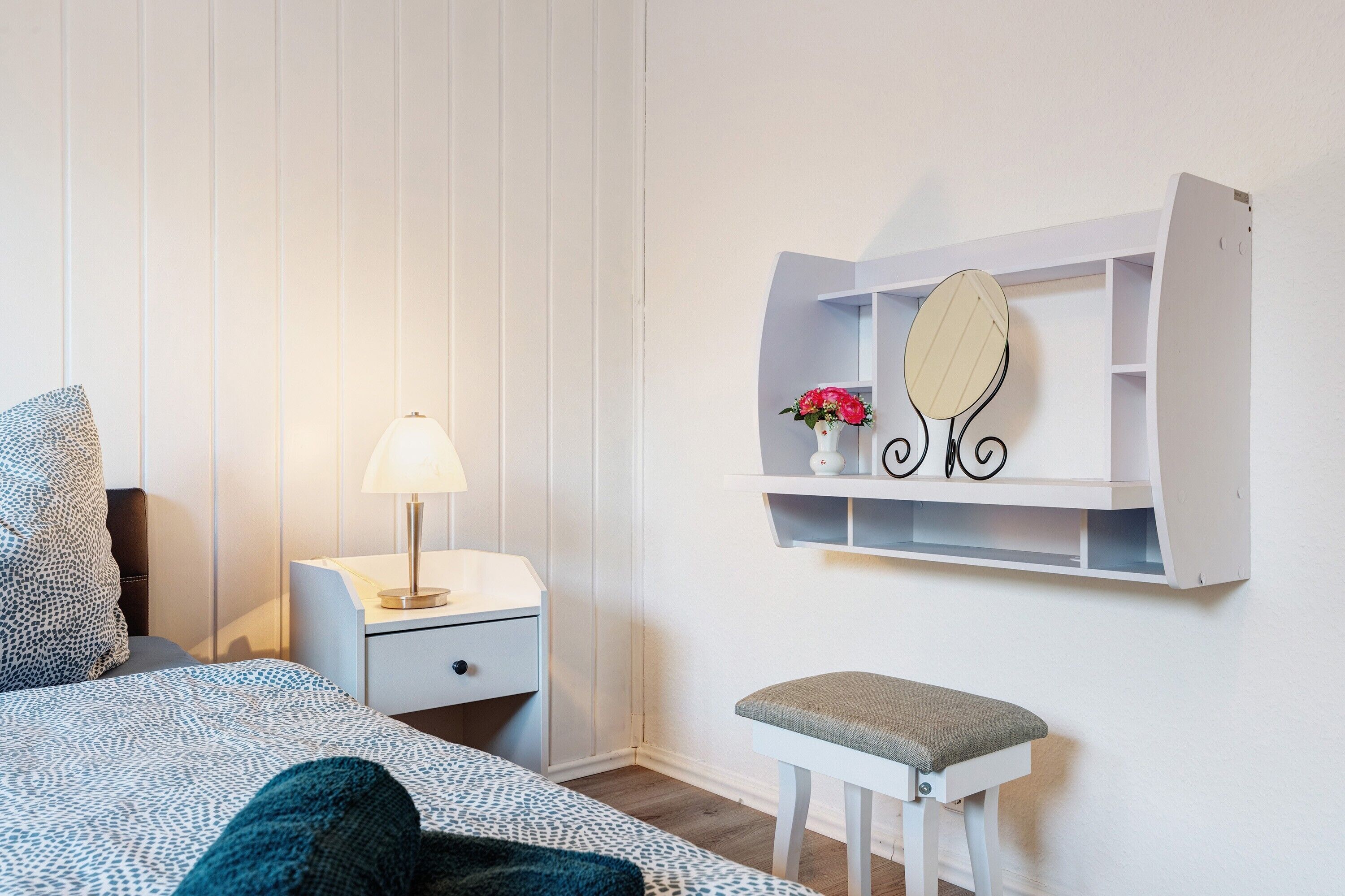 Un dormitorio con cama, mesita de noche, lámpara, tocador con espejo y un taburete pequeño.